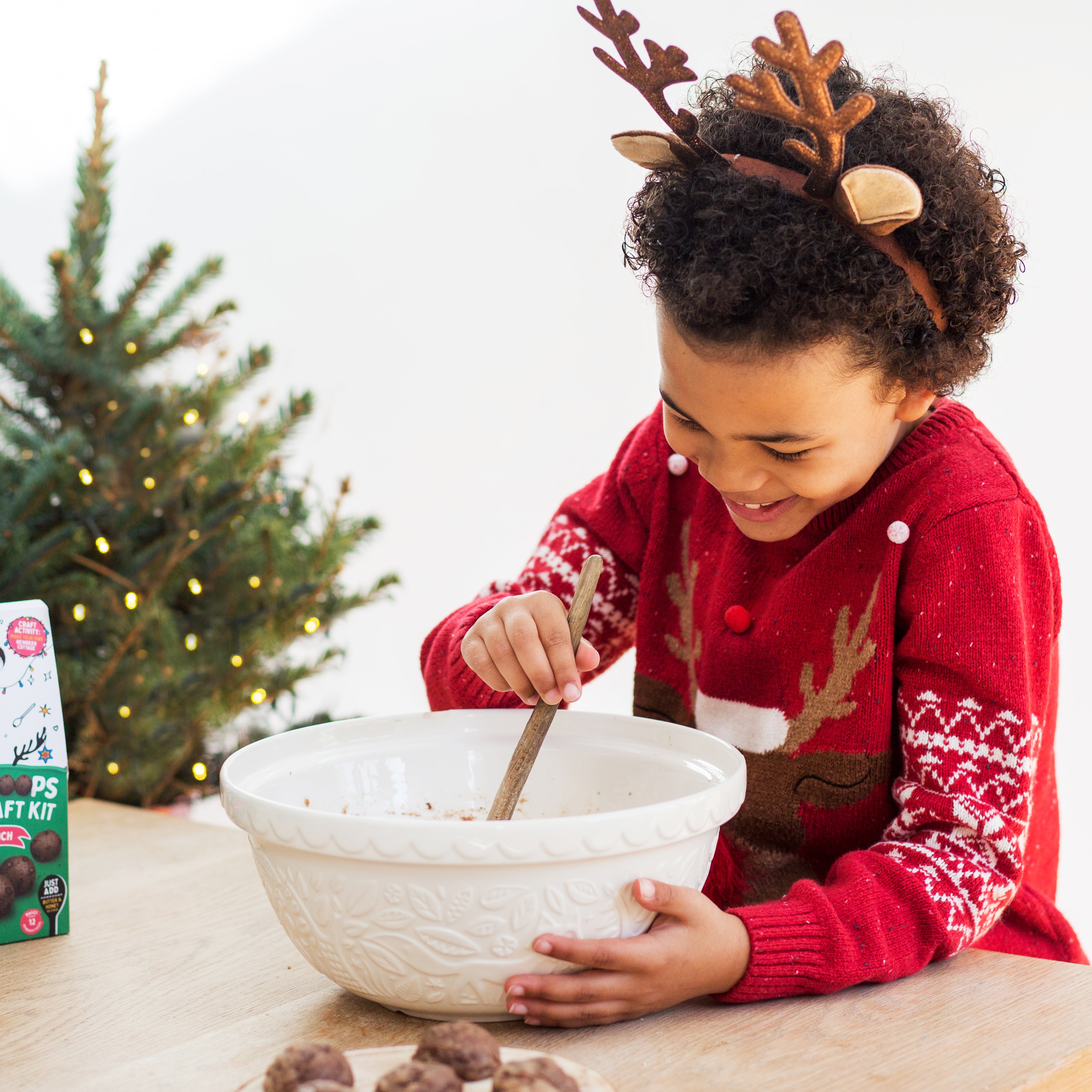 Child in reindeer sweater mixing dough in a bowl, with a small Christmas tree nearby.
