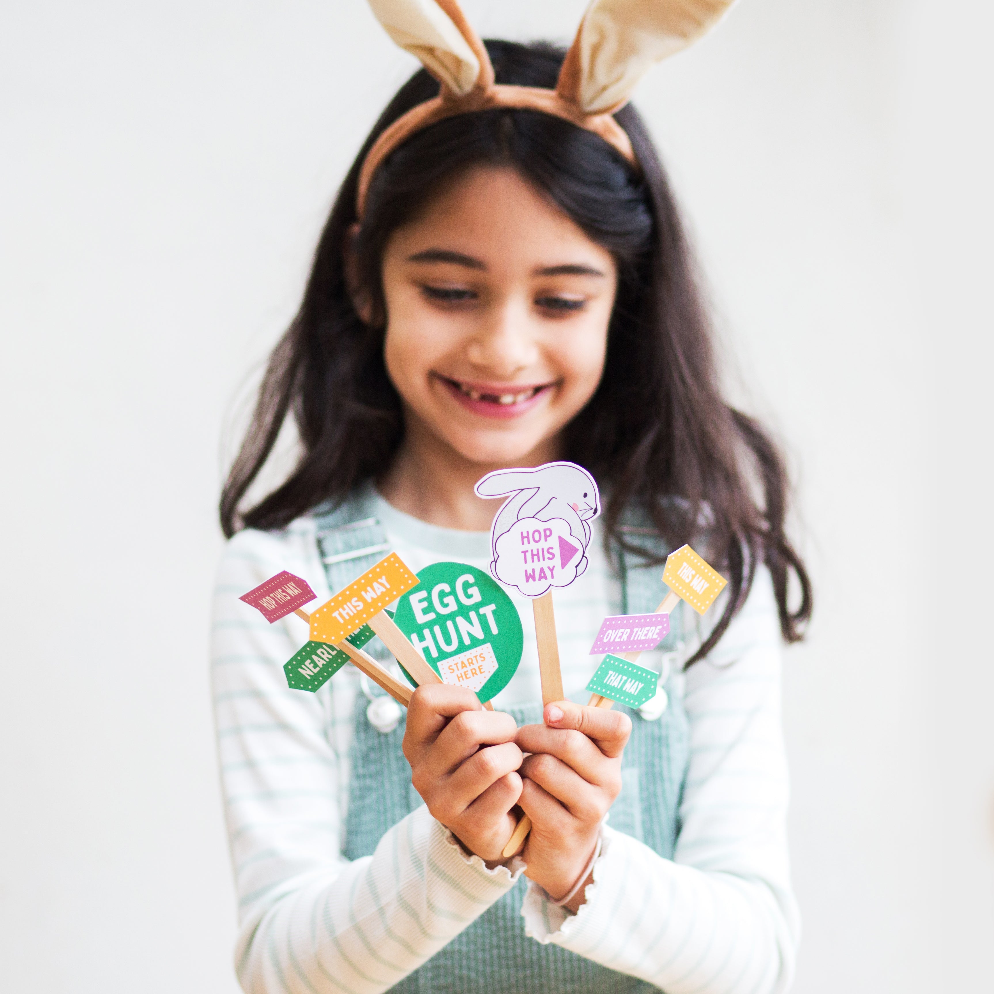 Girl with bunny ears holding Easter egg hunt signs, smiling.