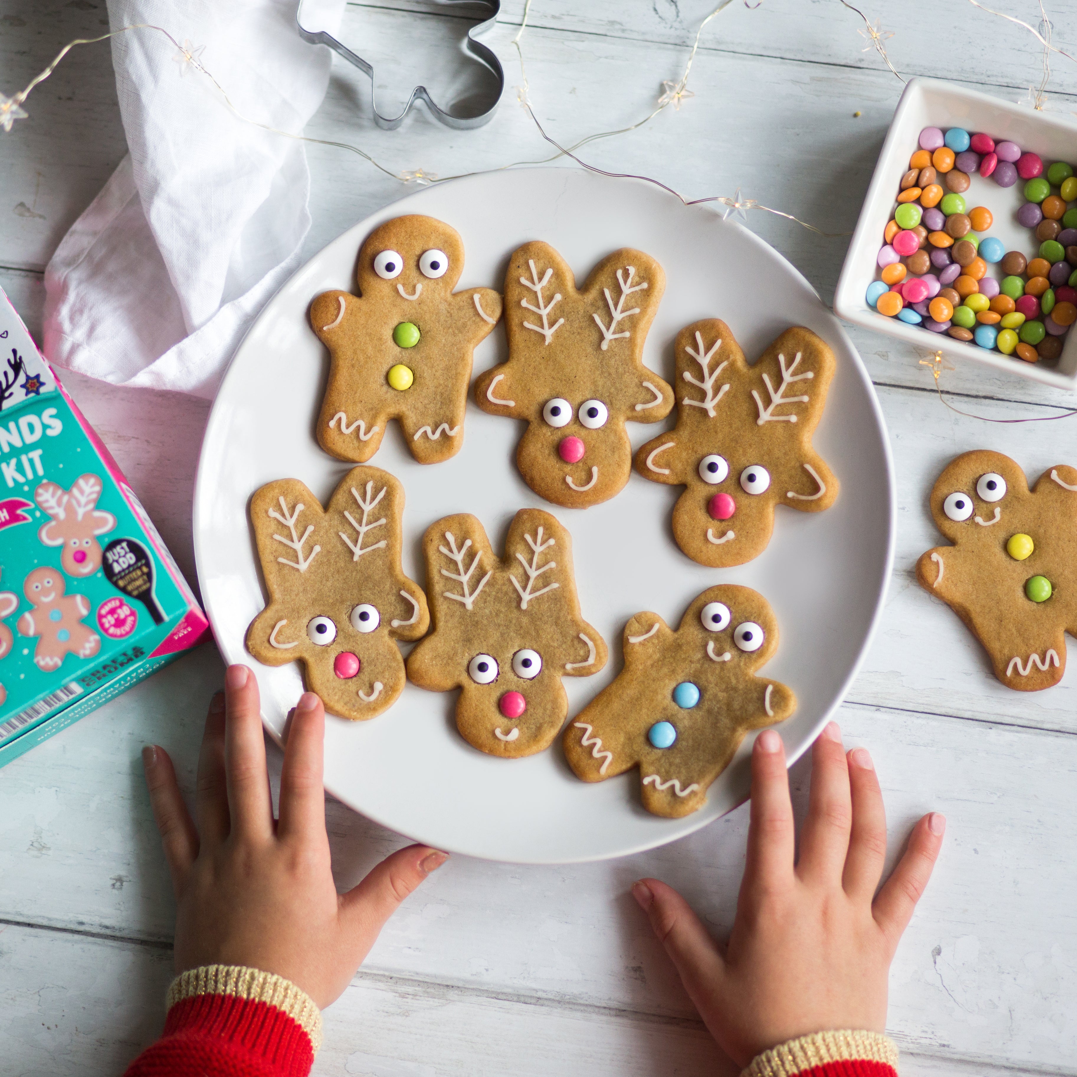 Child's hands reaching for a plate of decorated reindeer and gingerbread cookies with colorful candies on a wooden table.