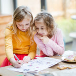 Two young girls drawing together at a table, surrounded by colorful papers and cookies.
