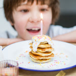 Child smiling at a stack of pancakes with sprinkles and a lit candle on top.
