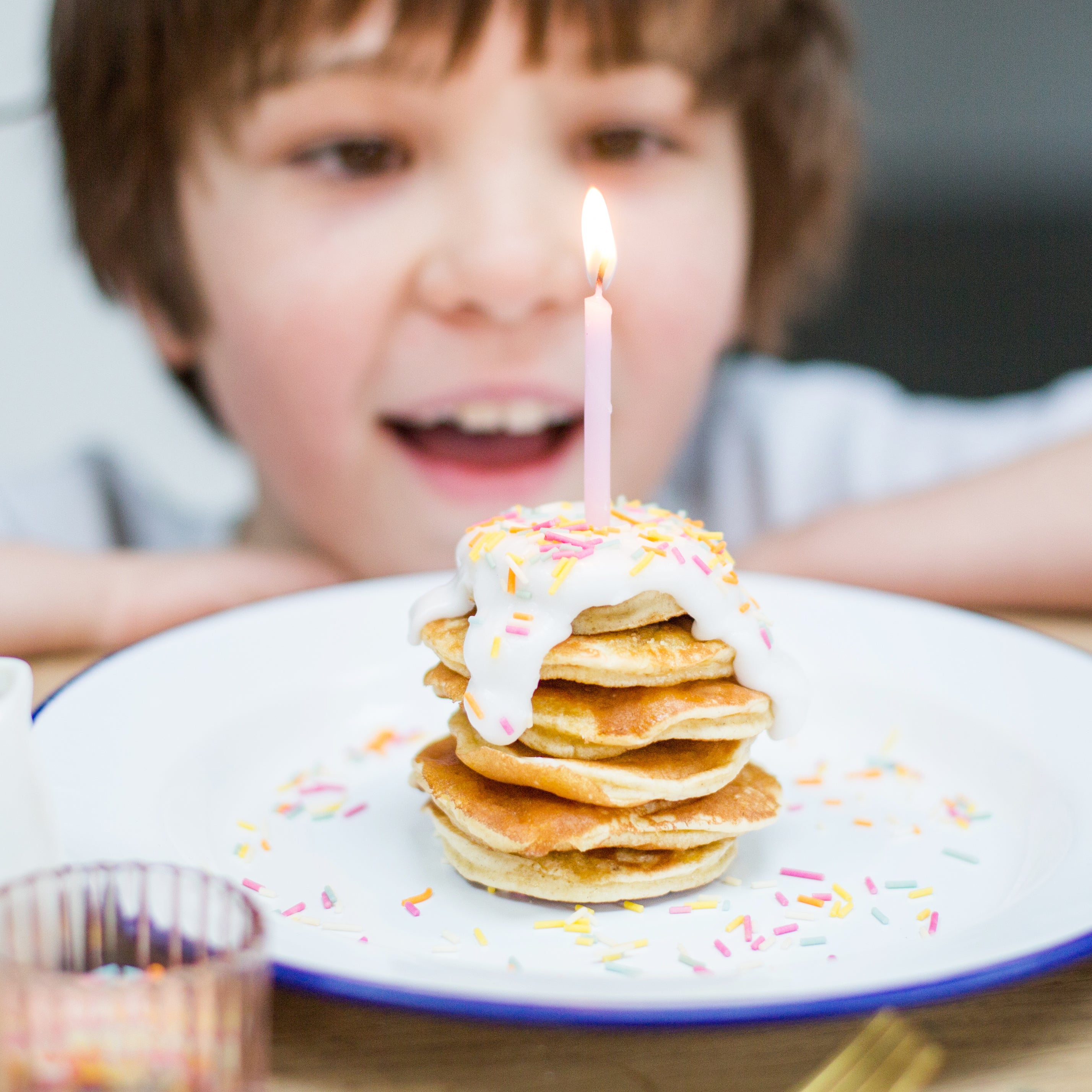 Child smiling at a stack of pancakes with sprinkles and a lit candle on top.