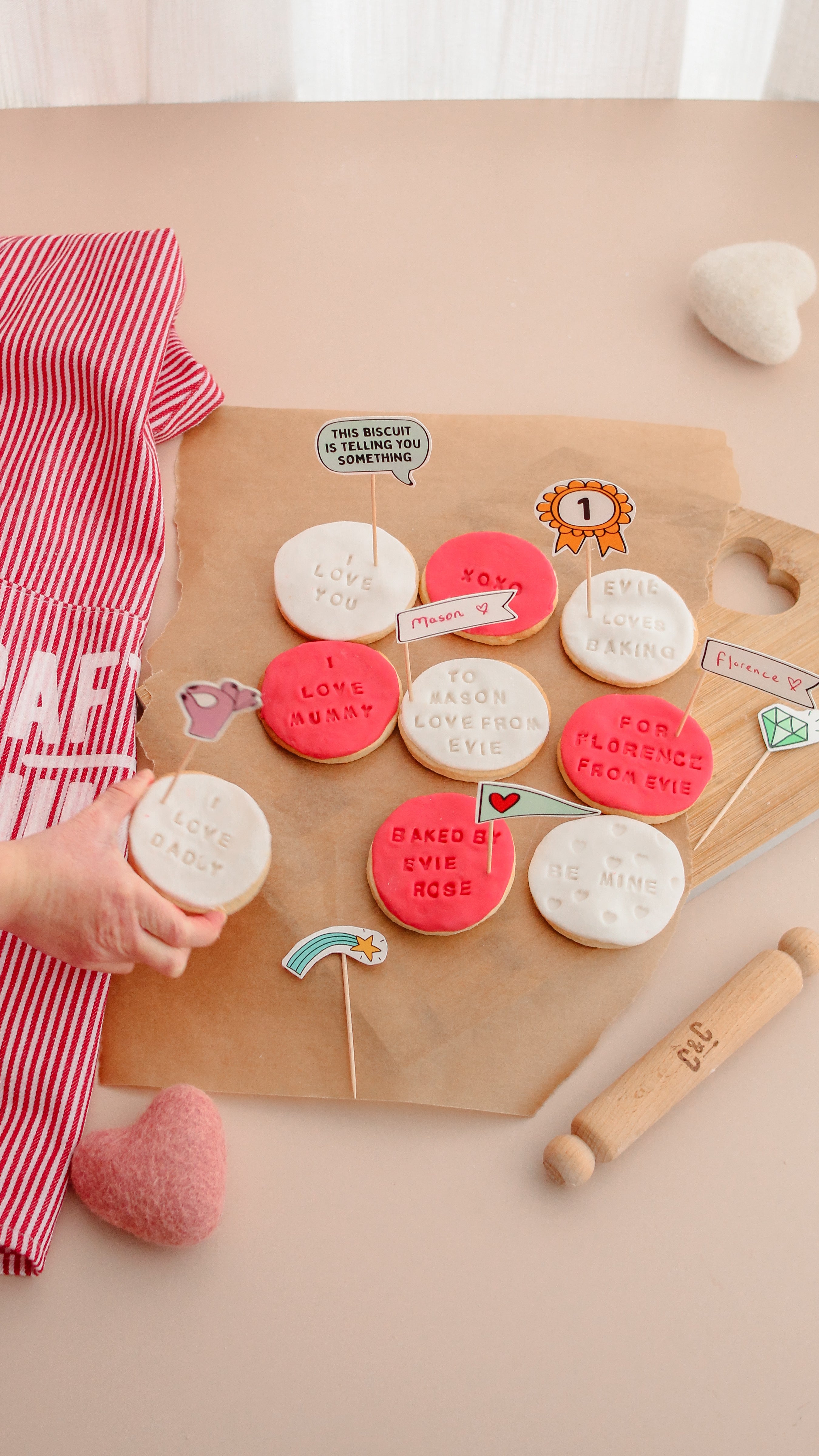 Decorated cookies with messages on a wooden board, child's hand reaching for one, surrounded by baking items.