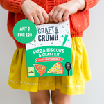 Child holding a Craft & Crumb pizza biscuits and craft kit, wearing a red sweater and yellow skirt.
