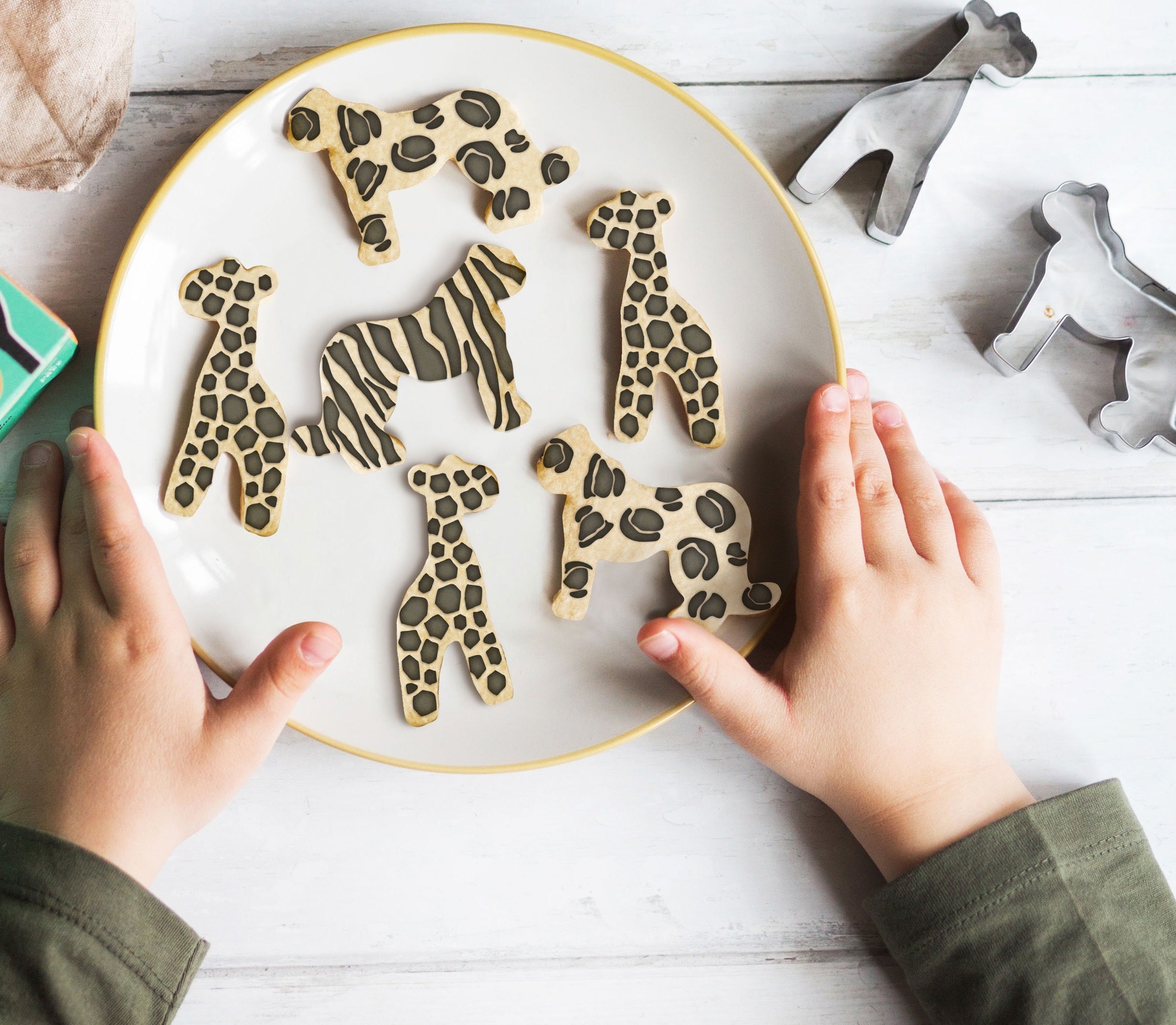 Child's hands holding a plate of safari animal cookies, including leopard, zebra, and giraffe designs.