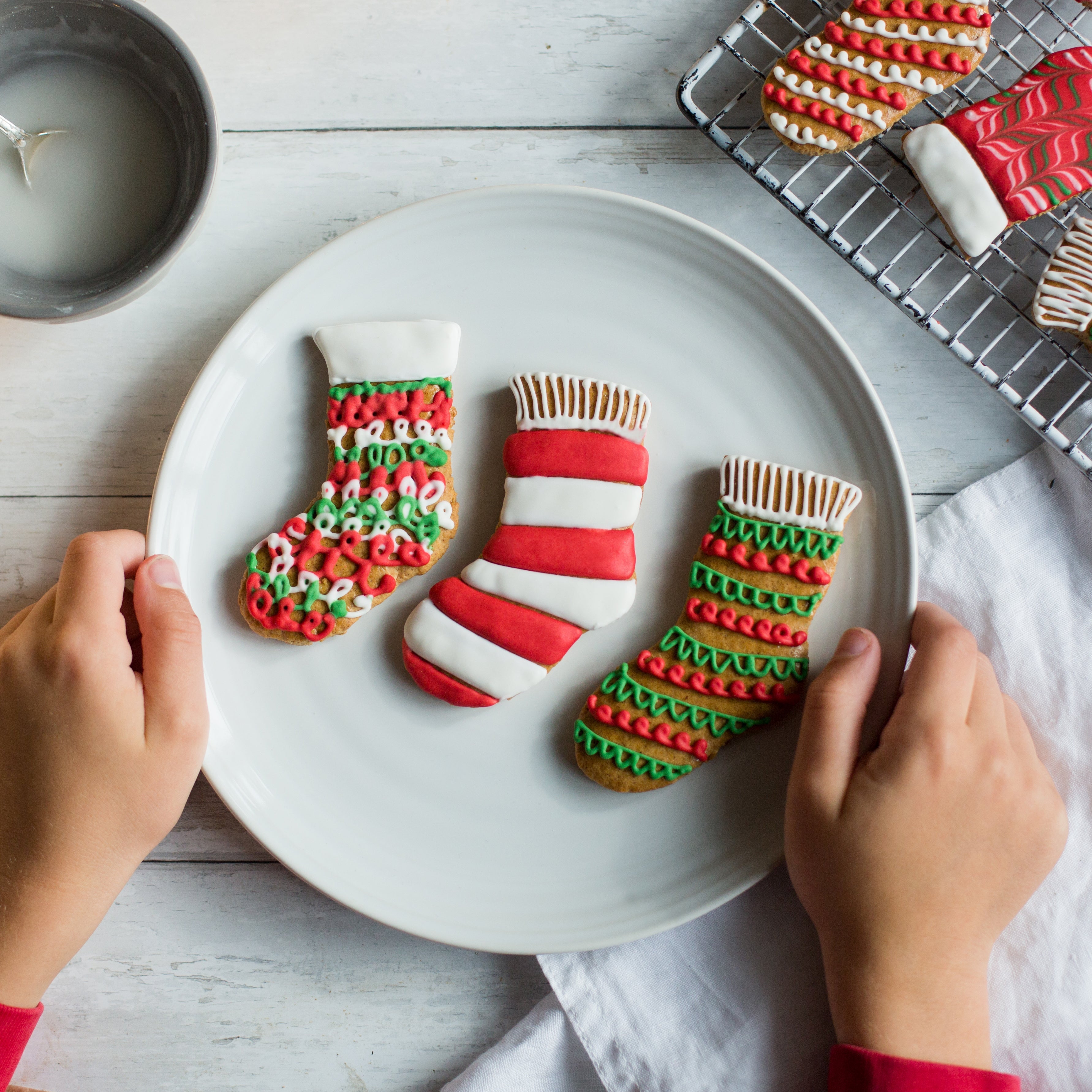 Hands holding a plate with three Christmas stocking cookies, decorated in red, green, and white icing.