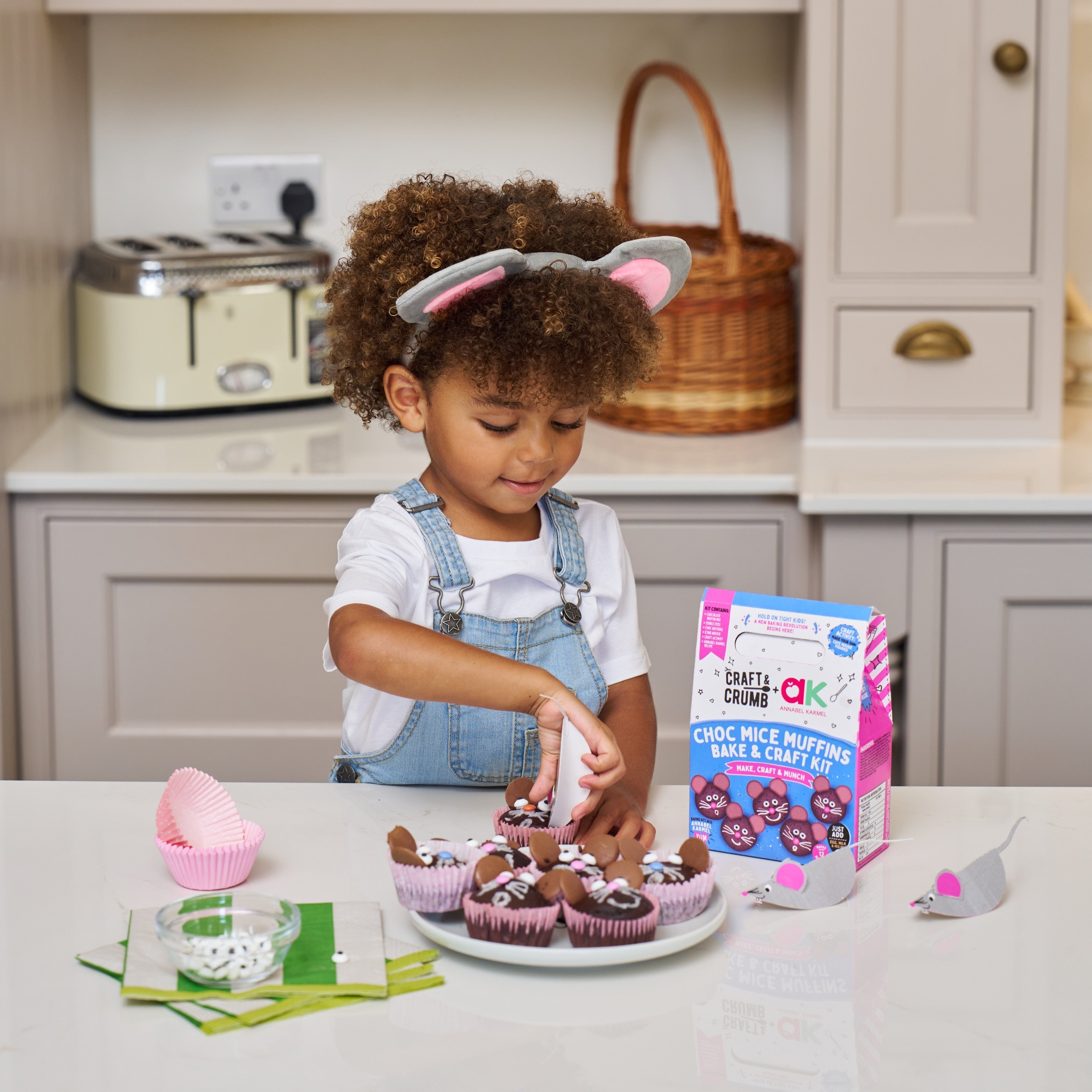 Child decorating cupcakes with chocolate icing in a kitchen, wearing mouse ears, next to a baking kit box.