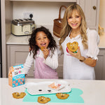 Child and woman smiling with teddy bear cookies in a kitchen, holding a cookie kit box.