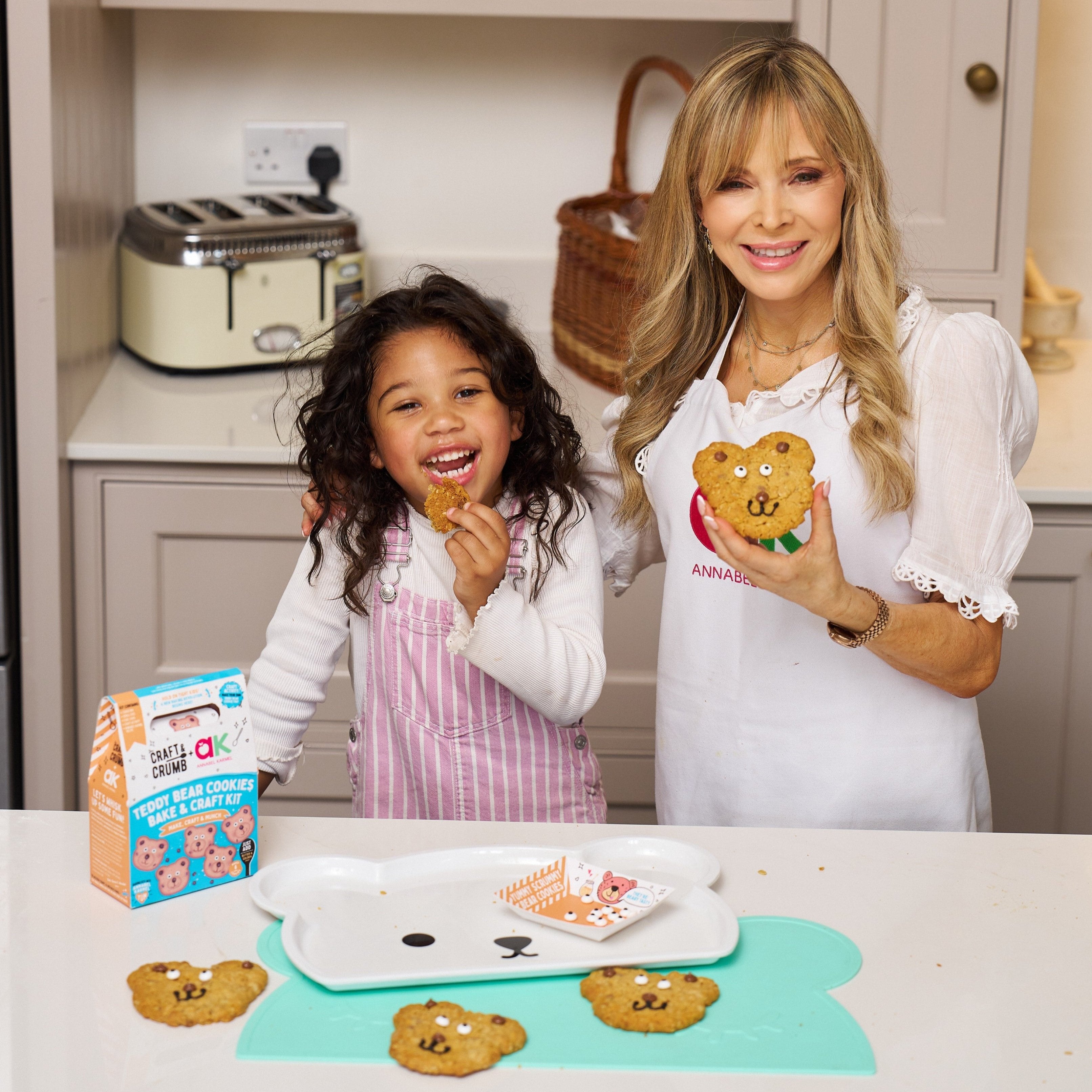Child and woman smiling with teddy bear cookies in a kitchen, holding a cookie kit box.