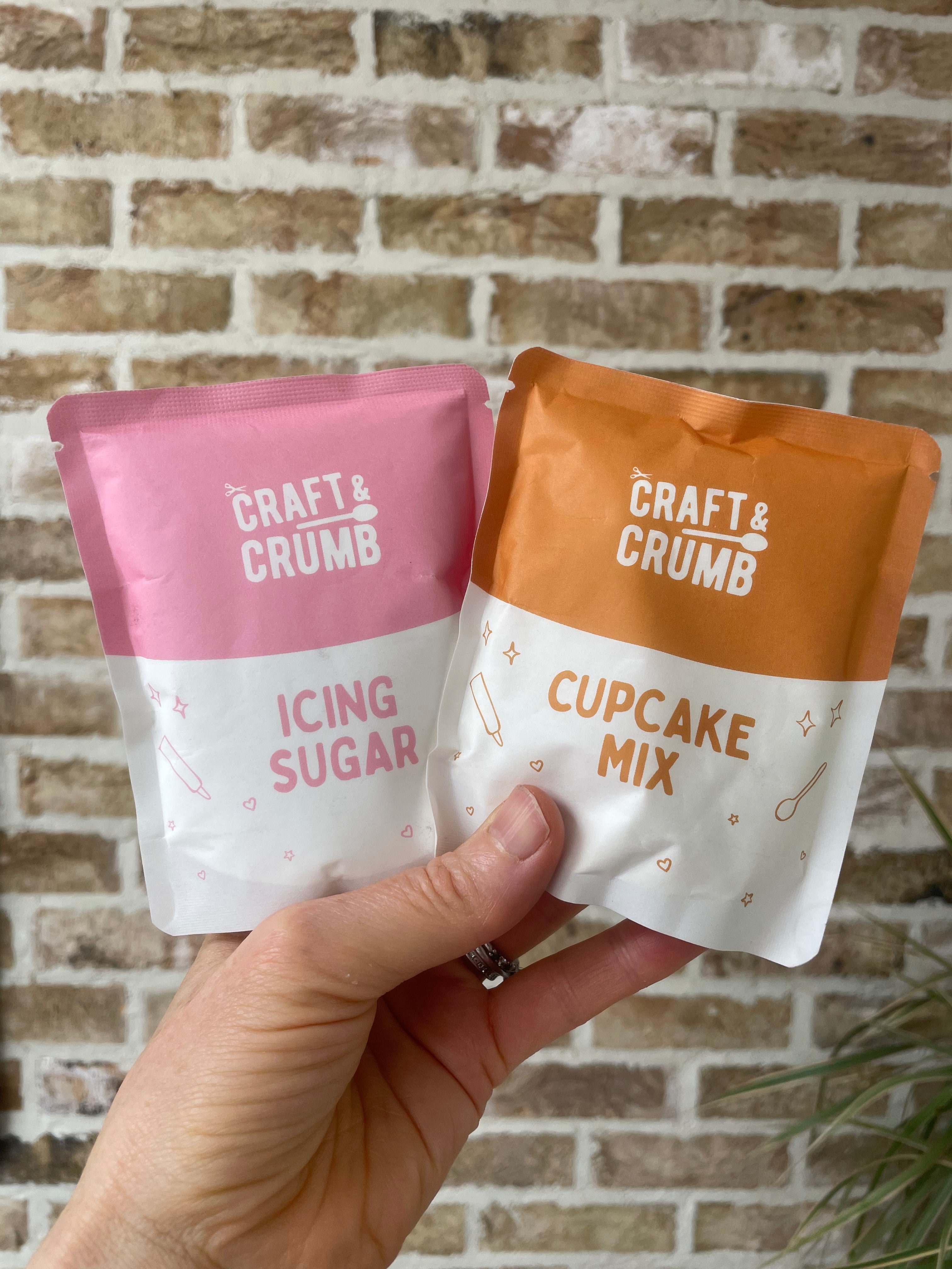Hand holding Craft & Crumb icing sugar and cupcake mix packets against a brick wall background.