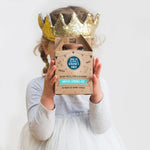 Child in a gold crown holding a "Sweetie Strings Kit" box, wearing a white dress against a plain background.
