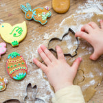 Child's hands using bunny cookie cutter on dough, surrounded by colorful Easter cookies on a wooden surface.