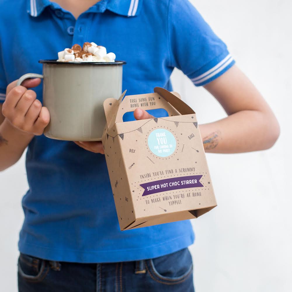 Person holding a mug of hot chocolate with marshmallows and a box labeled "Super Hot Choc Stirrer."