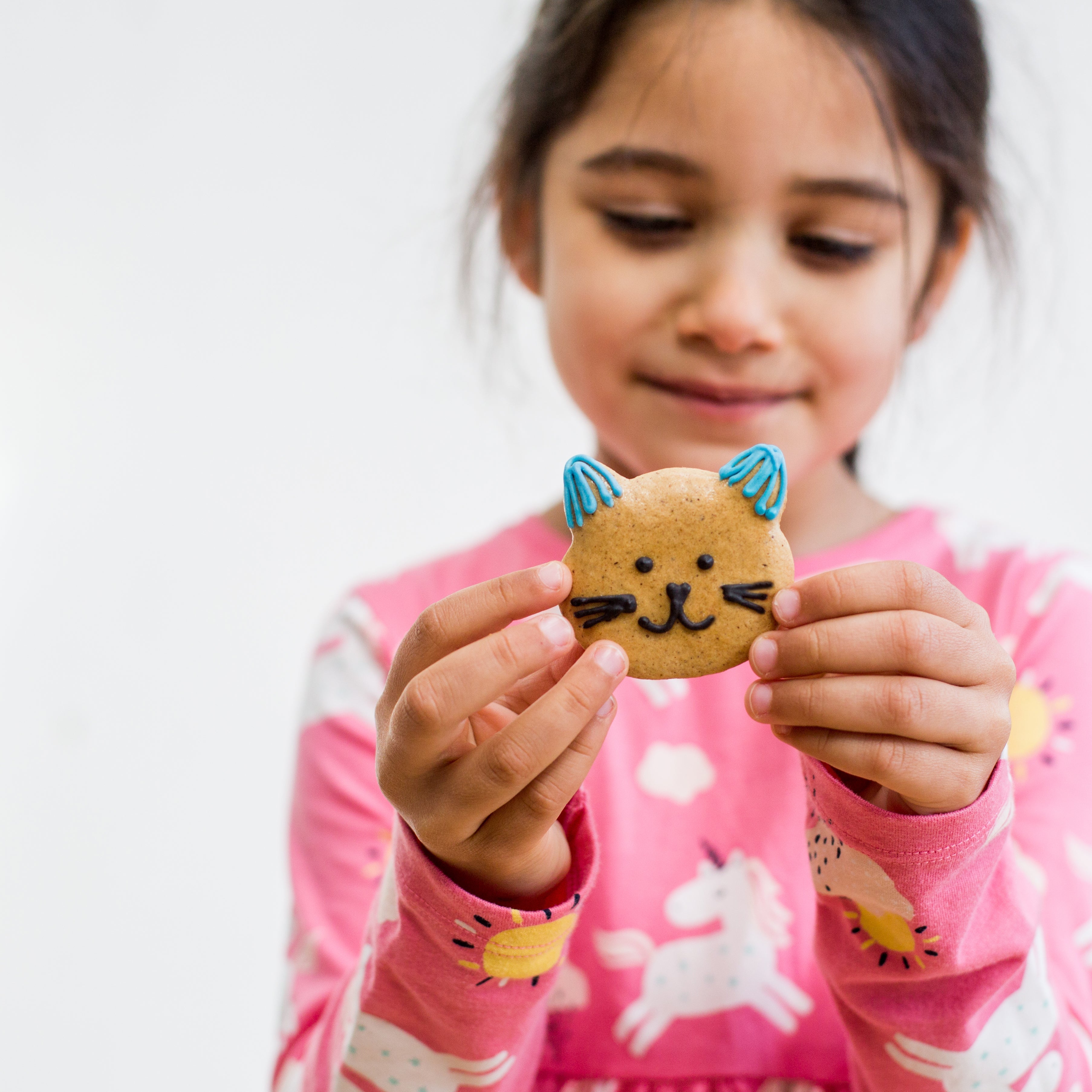 Girl in pink unicorn shirt holding a cat-shaped cookie with blue ears and whiskers, smiling gently.