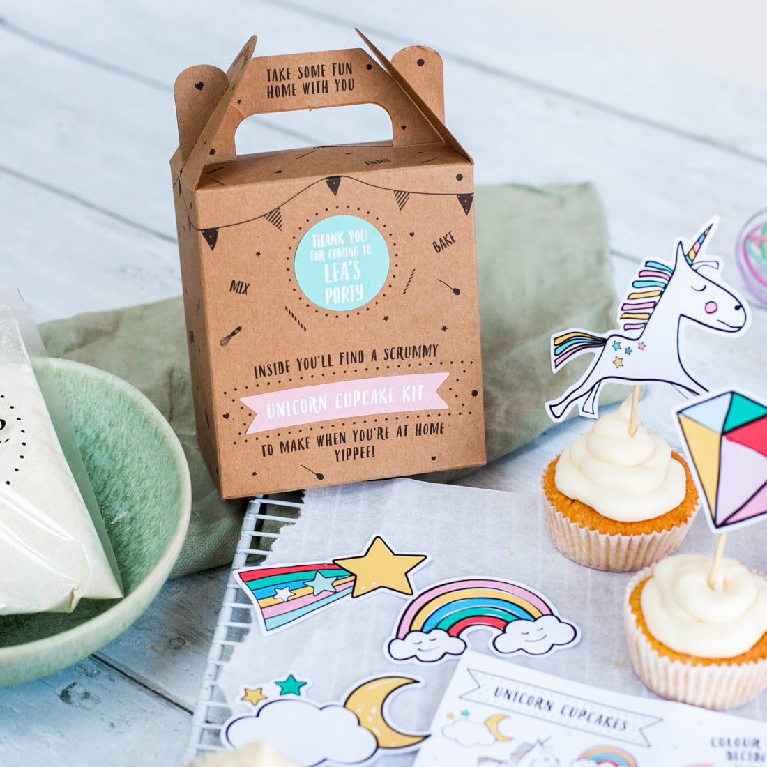 Unicorn-themed cupcake kit with decorations, cupcakes, and a party box on a table.