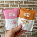 Hand holding Craft & Crumb icing sugar and cupcake mix packets against a brick wall background.