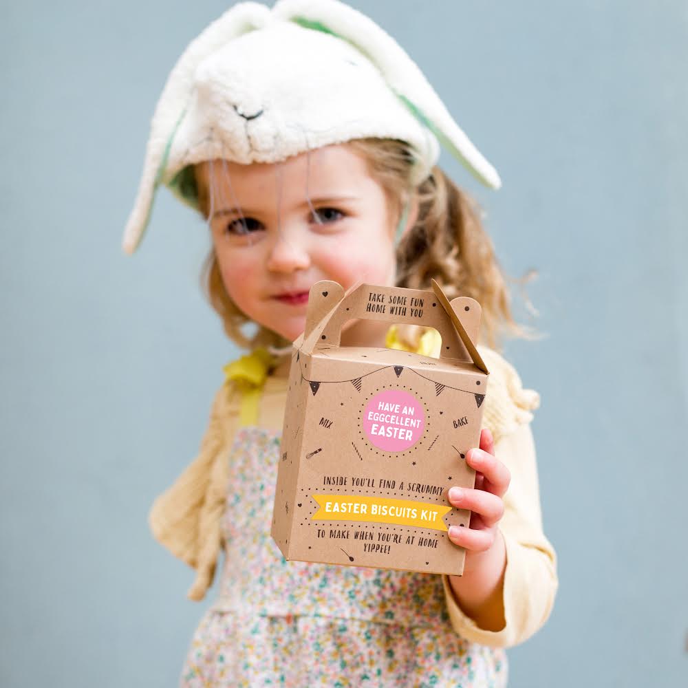 Child wearing bunny hat holding an Easter biscuits kit box, smiling against a plain background.