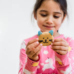 Girl in pink unicorn shirt holding a cat-shaped cookie with blue ears and whiskers, smiling gently.