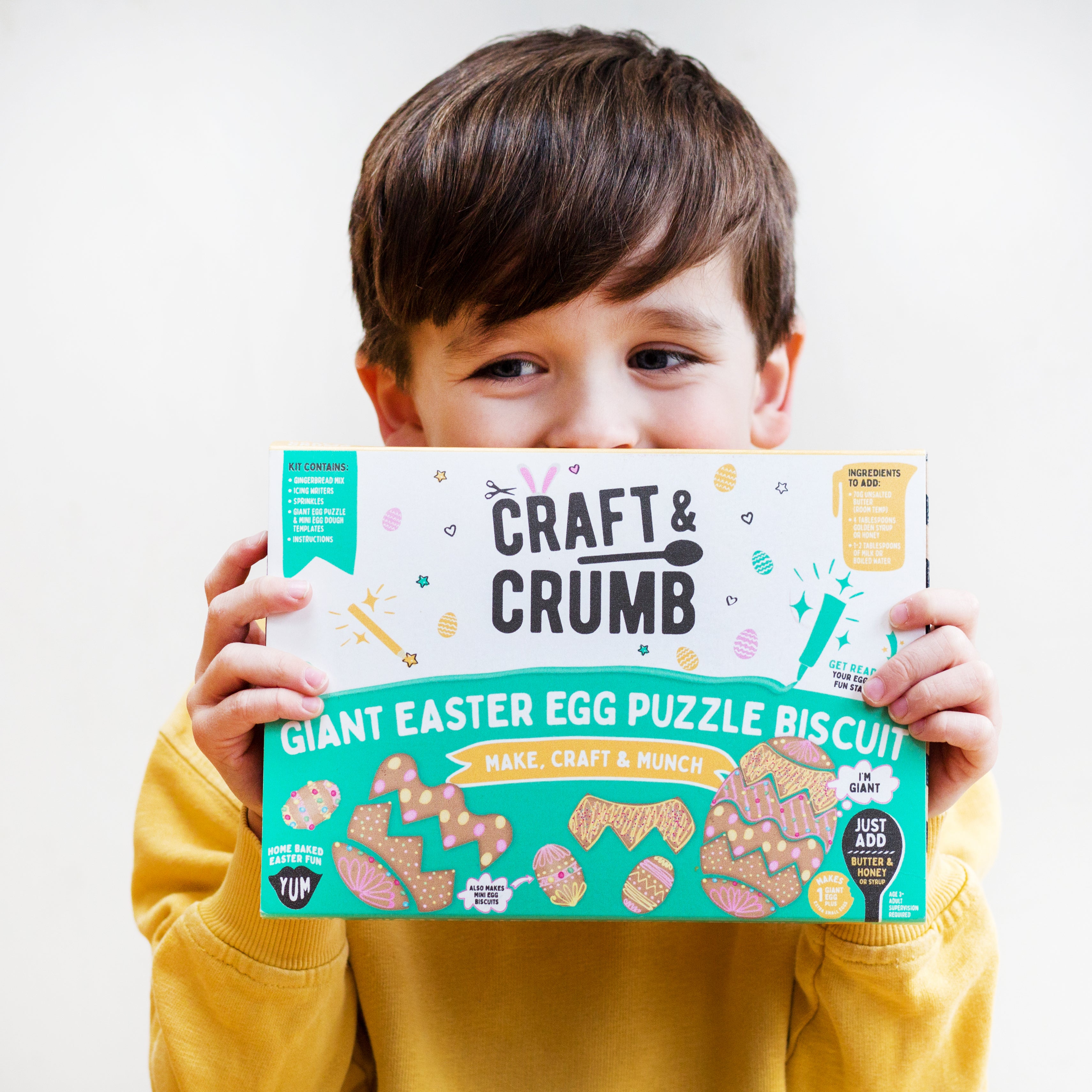 Child holding a Craft & Crumb Giant Easter Egg Puzzle Biscuit box, smiling, wearing a yellow sweater.