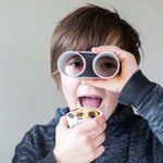 Child holding a cupcake and looking through homemade binoculars made from paper rolls, wearing a dark hoodie.