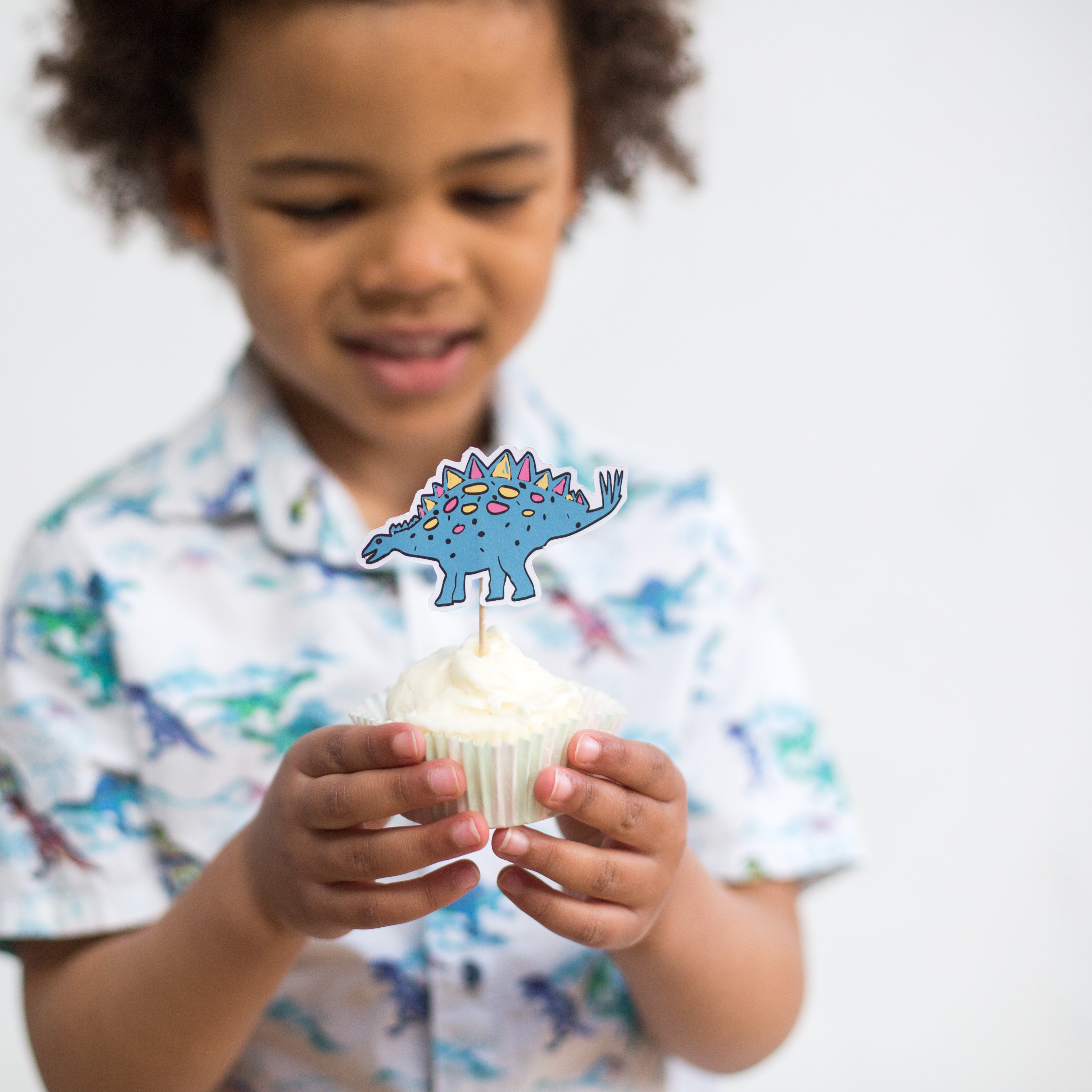 Child holding a cupcake with a blue dinosaur topper, wearing a dinosaur-patterned shirt.