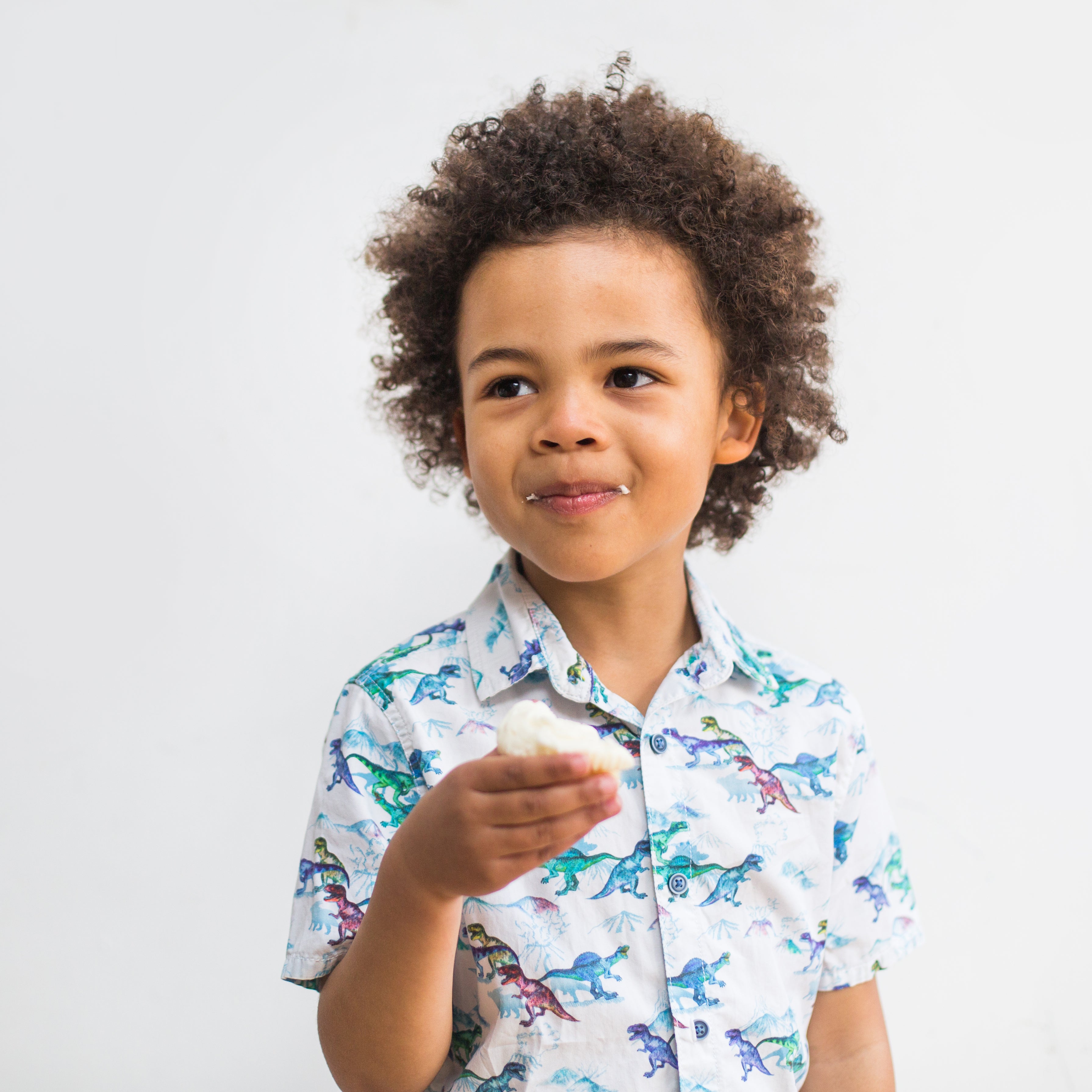 Young child with curly hair eating a treat, wearing a dinosaur-patterned shirt, against a plain white background.