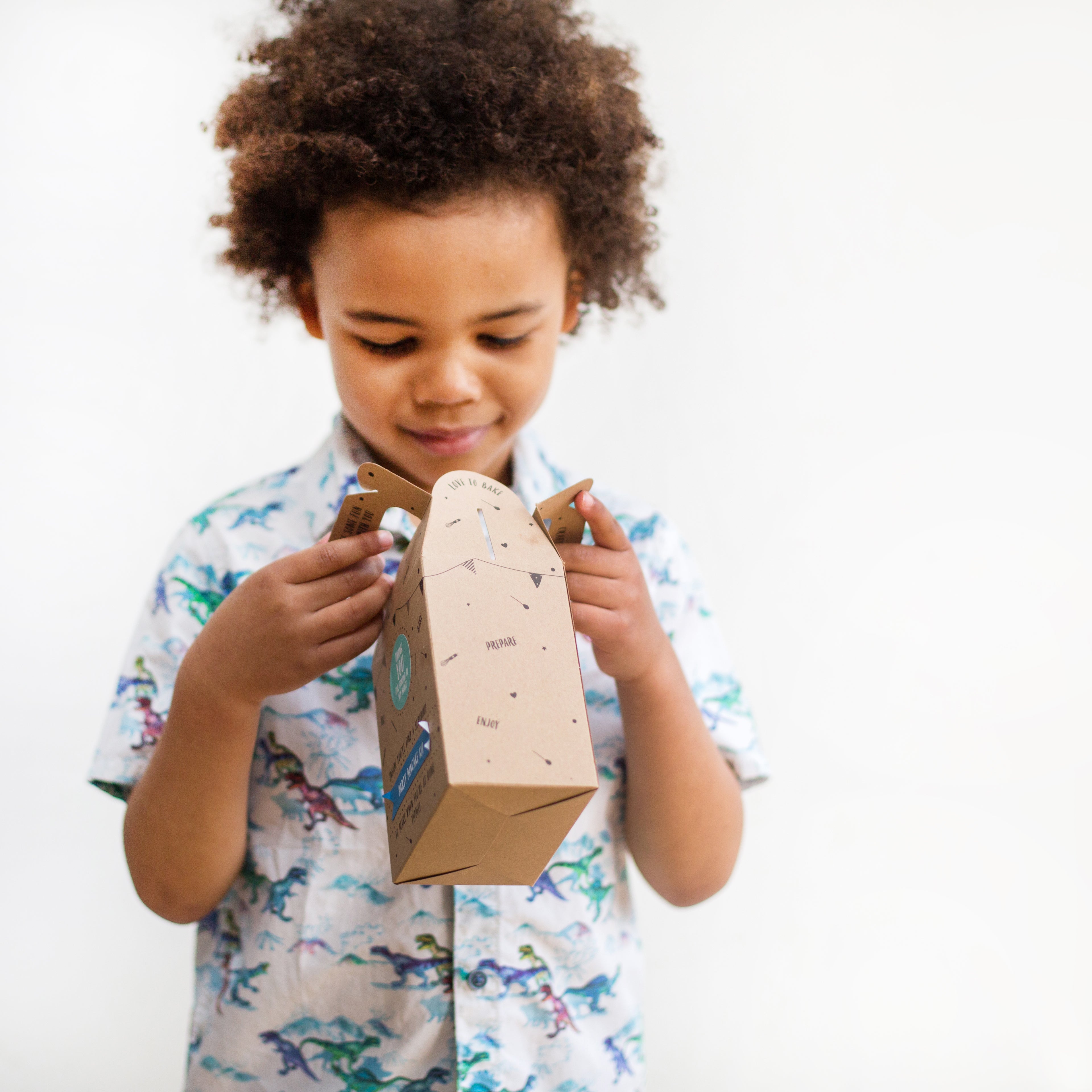 Young child in a dinosaur shirt holding a cardboard craft box, looking down with a curious expression.
