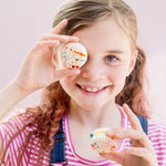 Girl smiling, holding colorful speckled macarons, one over her eye. Wearing a pink striped shirt and denim overalls.