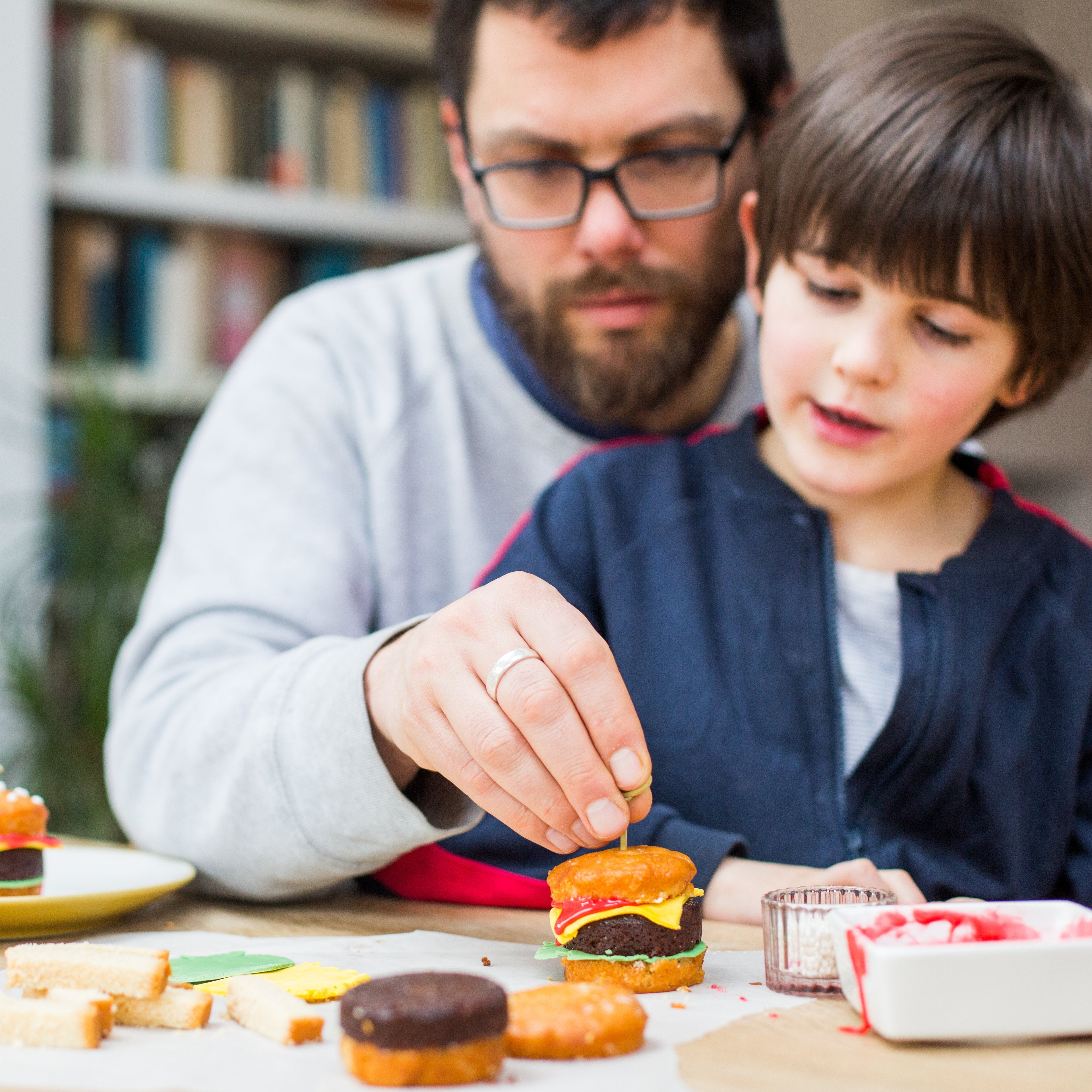 Adult and child assembling mini burger-shaped cakes at a table, with bookshelves in the background.