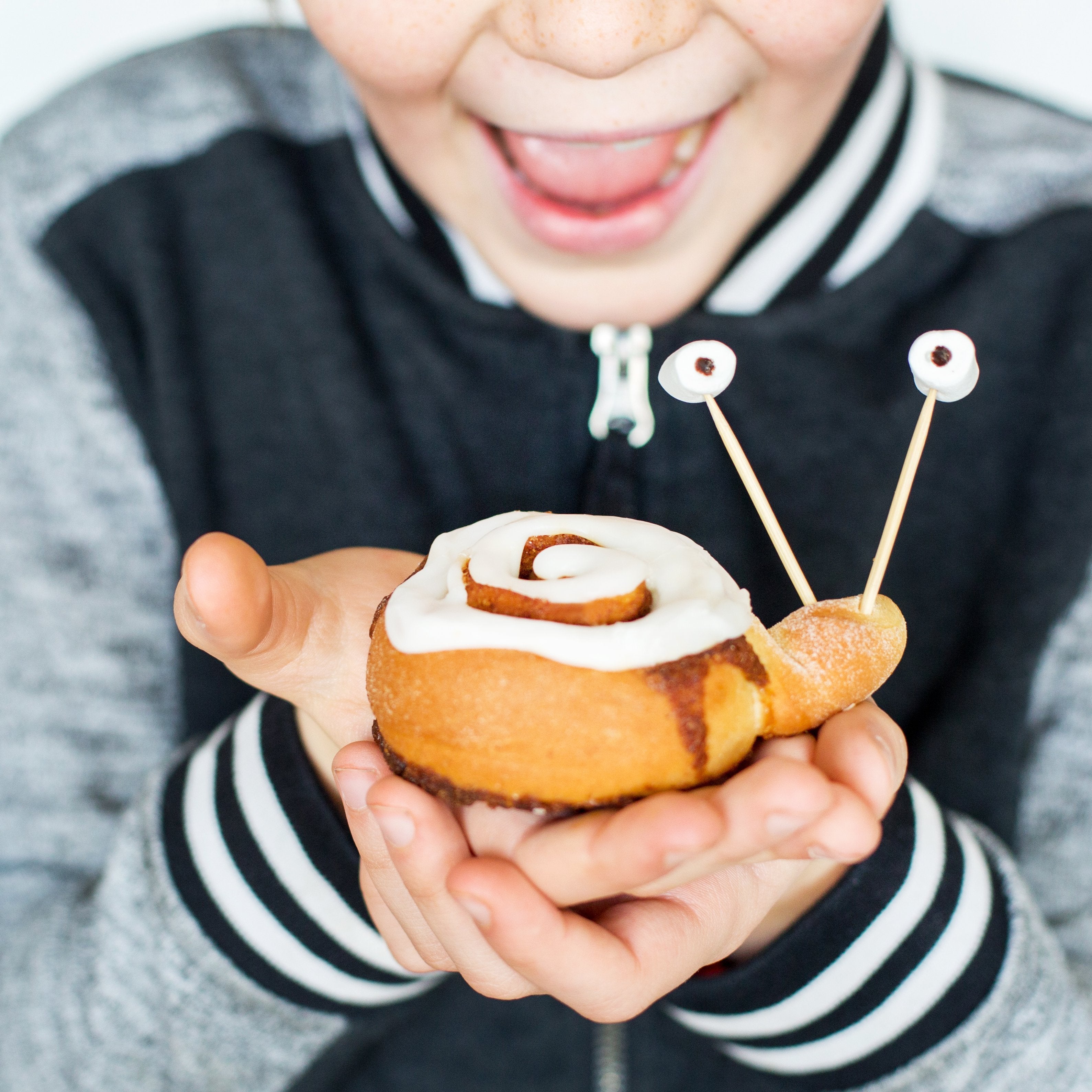 Child holding a cinnamon roll shaped like a snail with icing and candy eyes, smiling excitedly.