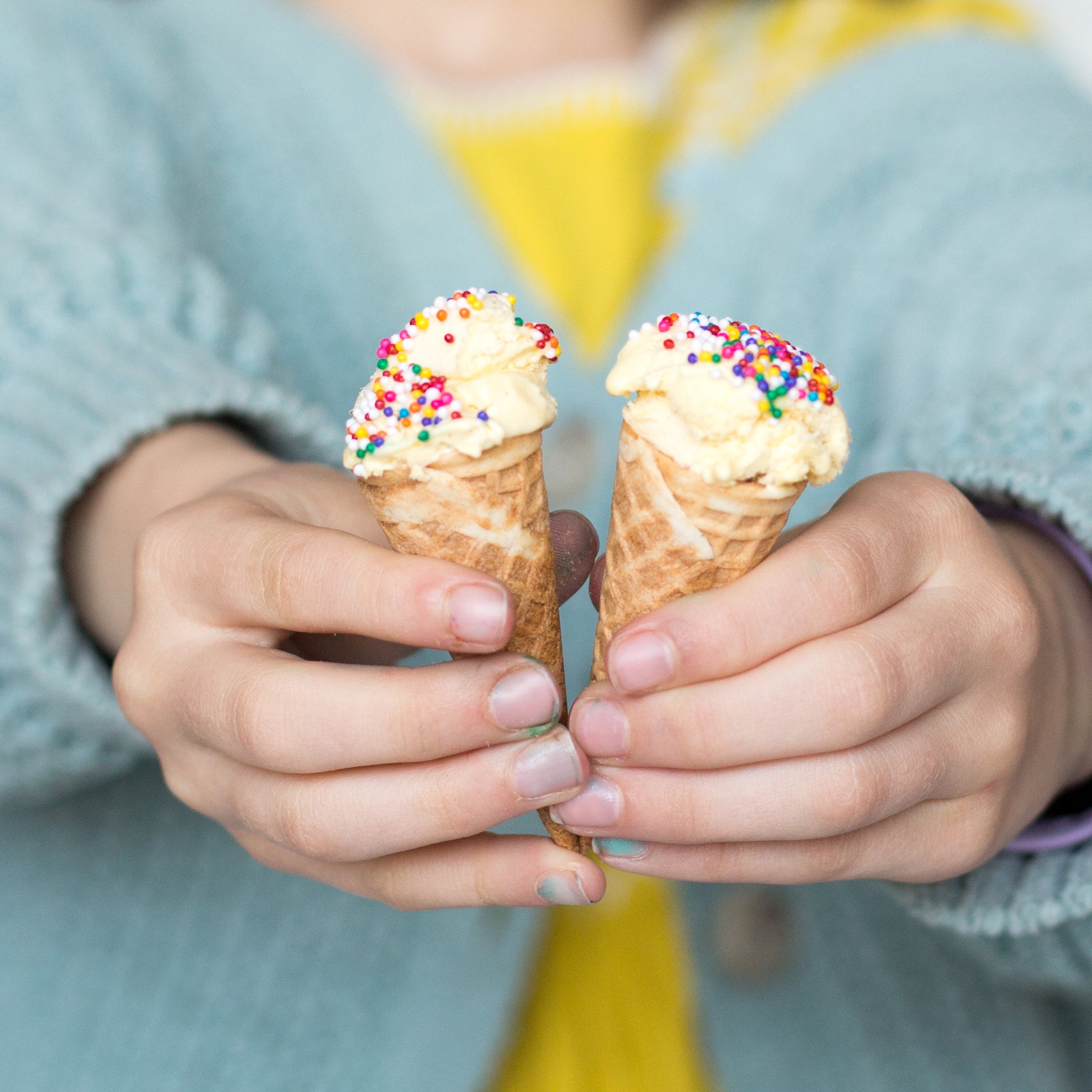 Child holding two small ice cream cones with sprinkles, wearing a blue sweater and yellow shirt.