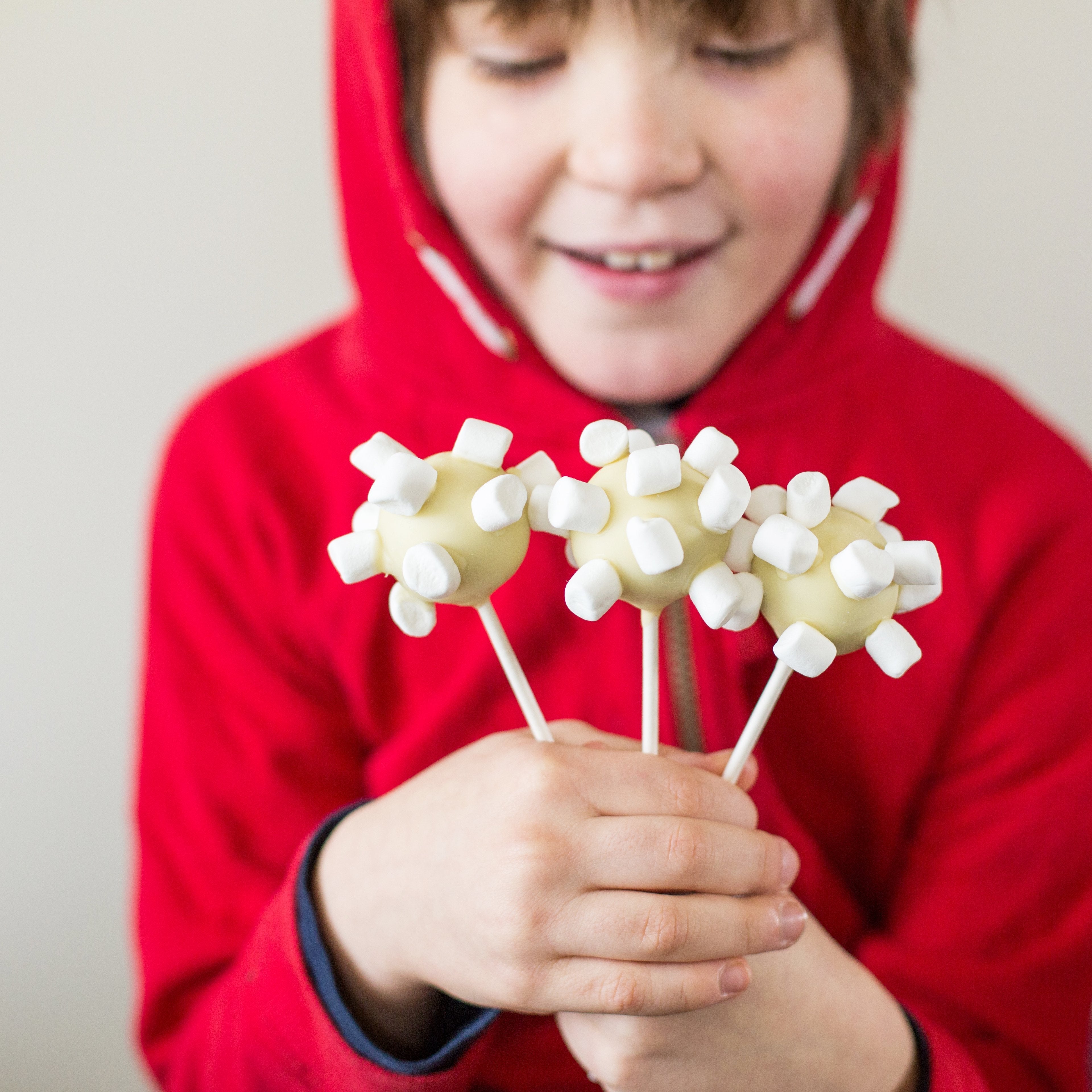 Child in red hoodie holding three cake pops decorated with white marshmallows, resembling a virus shape.