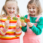 Two smiling children holding colorful, decorated cookies shaped like a flower, carrot, pineapple, and snail.