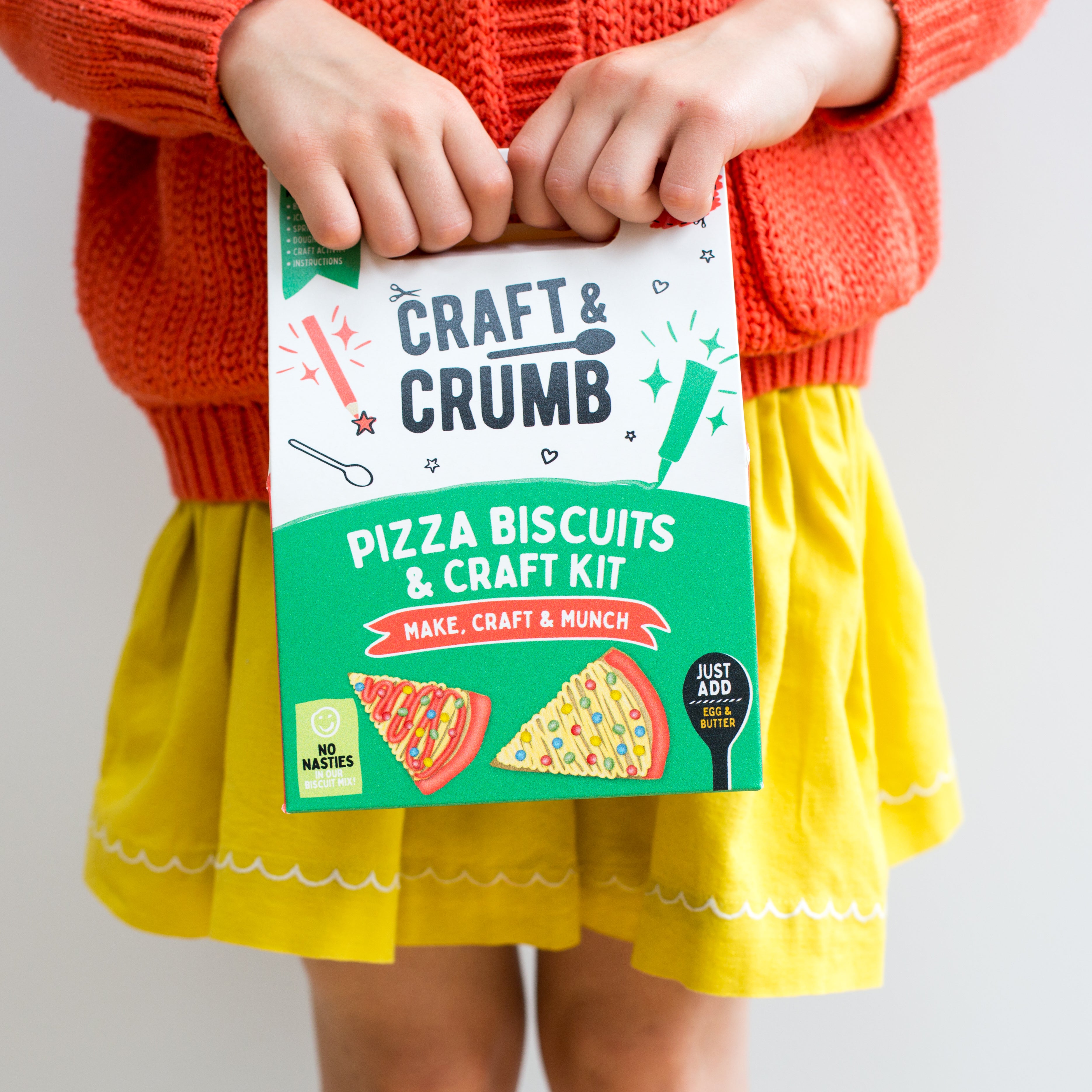 Child holding a "Craft & Crumb" pizza biscuits and craft kit box, wearing a red sweater and yellow skirt.