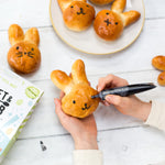 Hands decorating bunny-shaped bread rolls with chocolate icing on a white table.