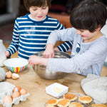 Two children baking, mixing ingredients in a bowl, surrounded by eggs, milk, and pancakes on a wooden table.