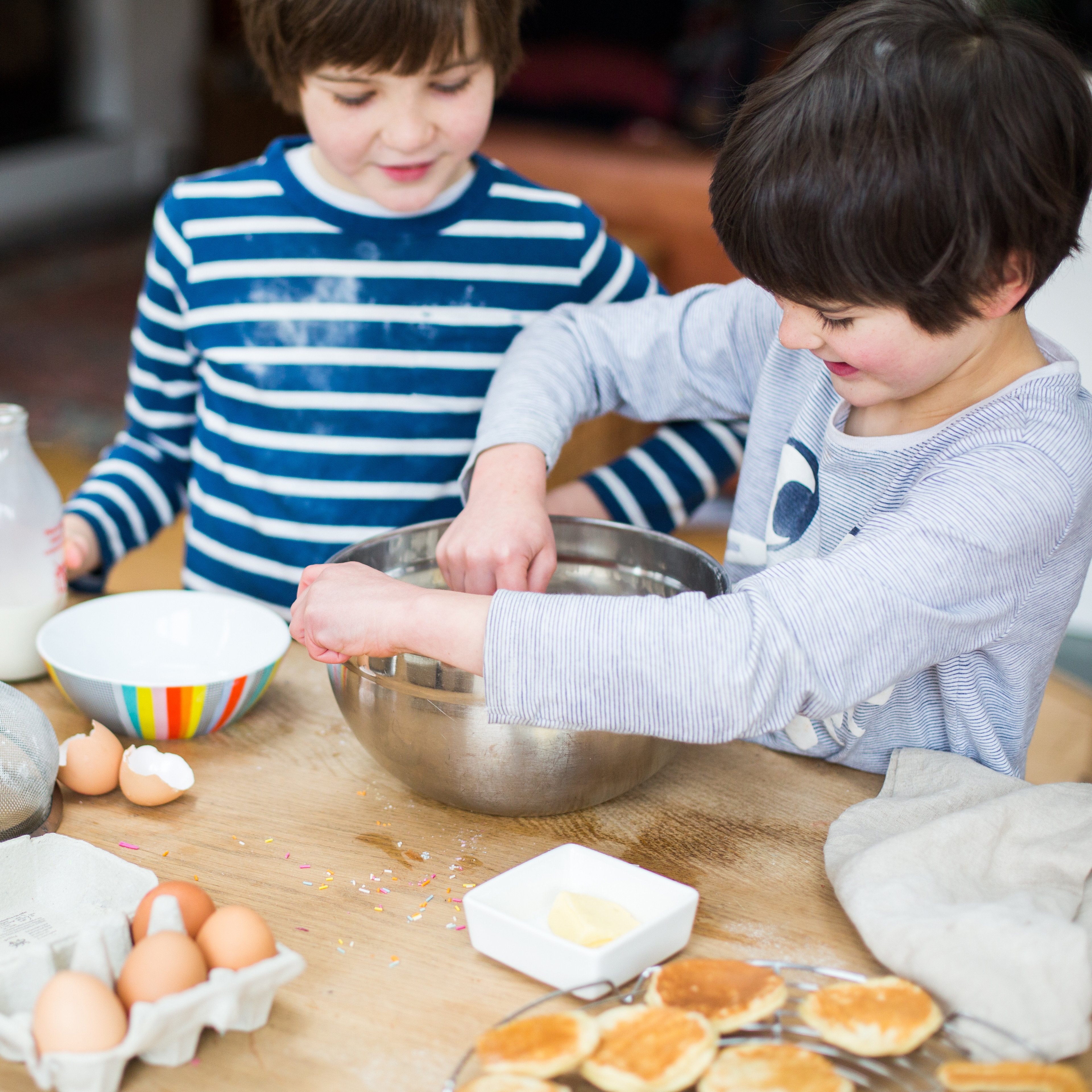 Two children baking, mixing ingredients in a bowl, surrounded by eggs, milk, and pancakes on a wooden table.