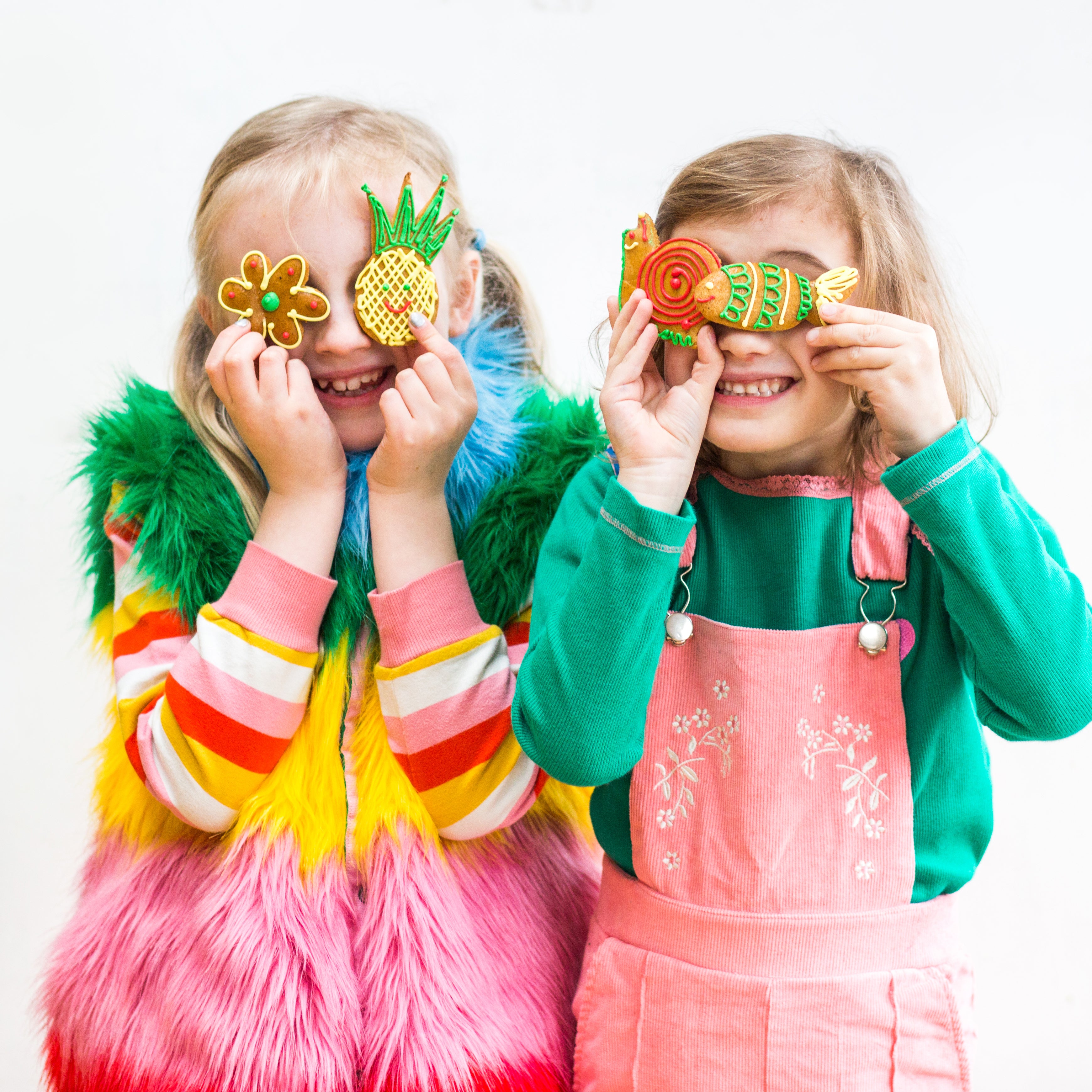 Two children smiling, holding colorful fruit-shaped cookies over their eyes, wearing vibrant outfits.