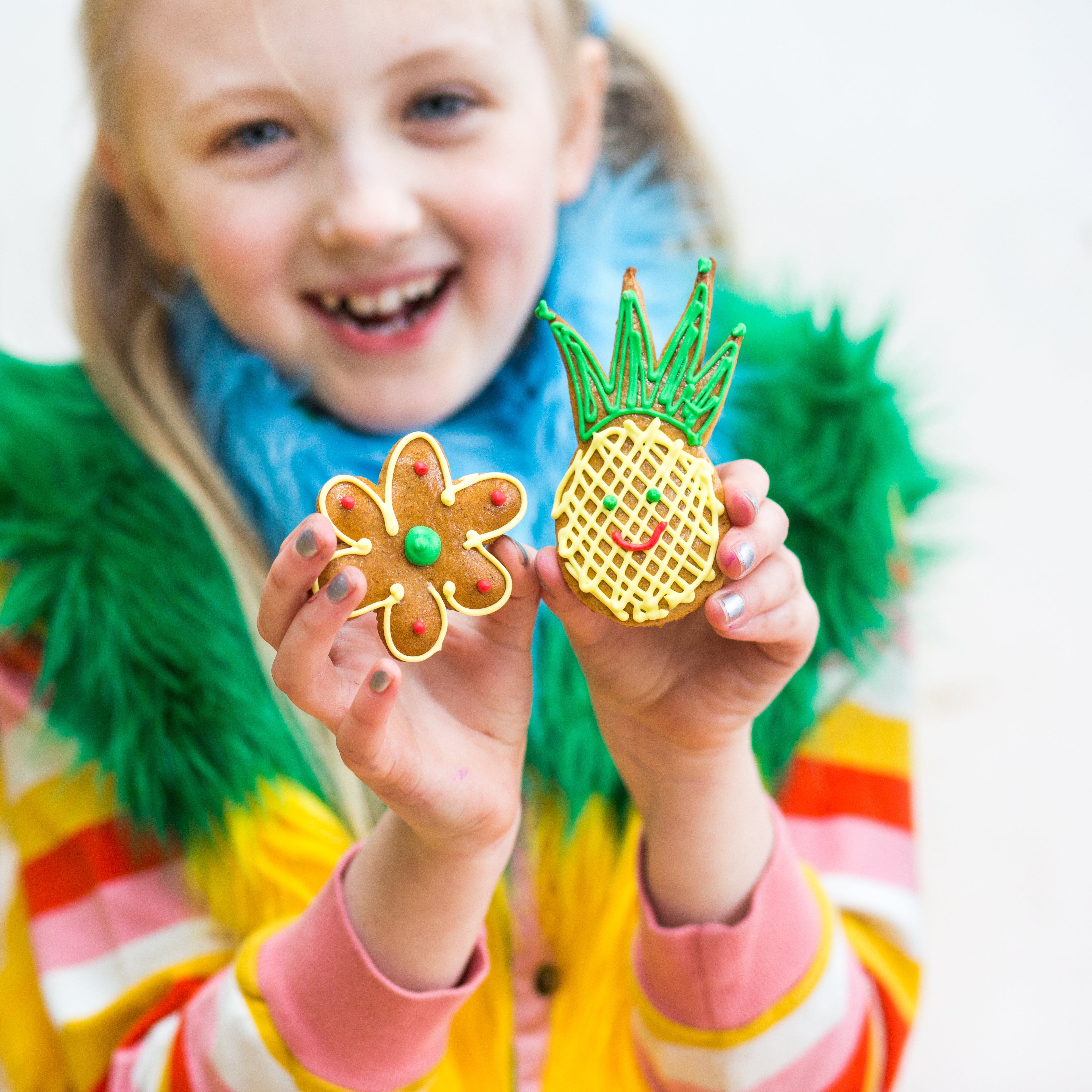 Child holding a flower-shaped cookie and a pineapple-shaped cookie with green icing, wearing colorful clothes and smiling.