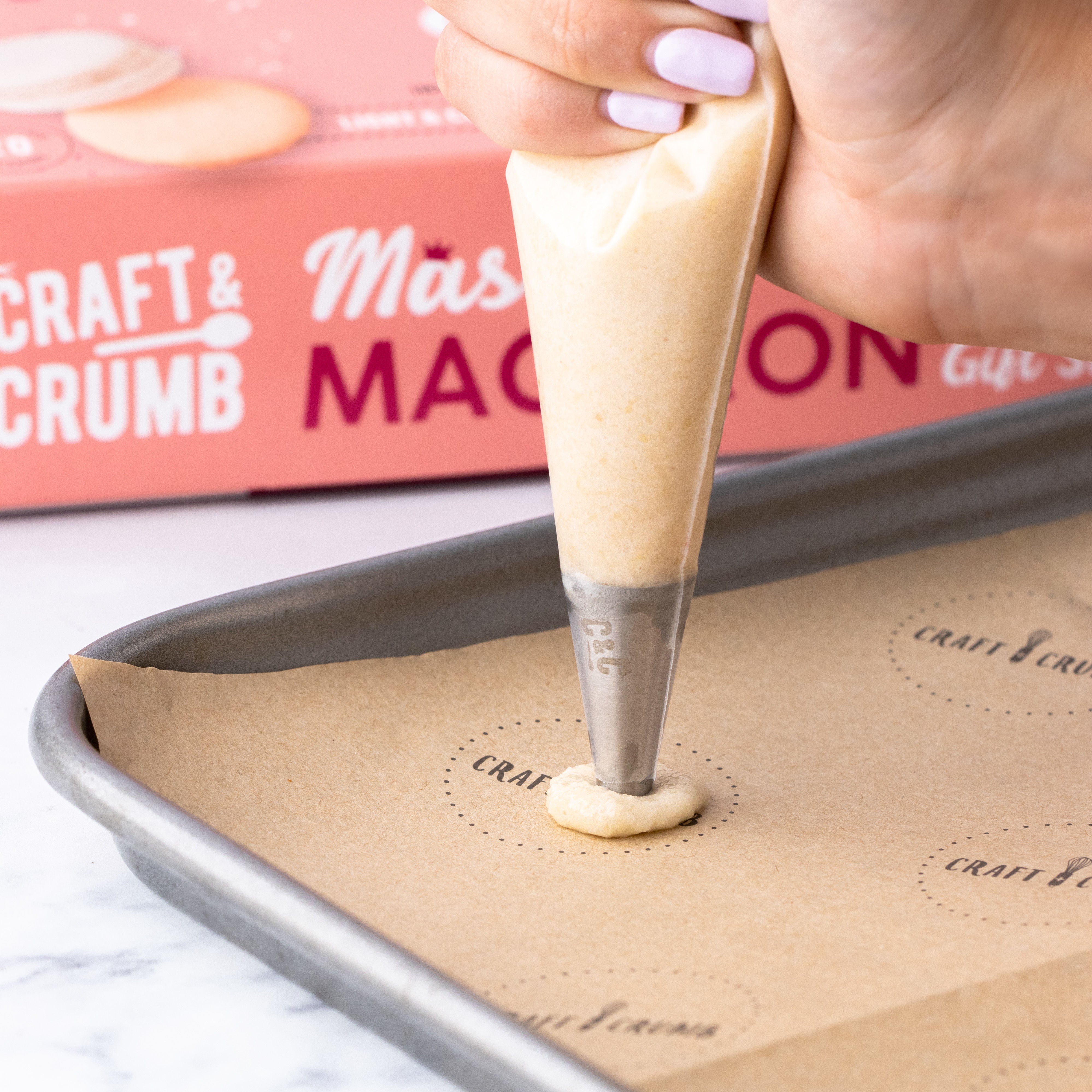Hand piping macaron batter onto a baking sheet with a Craft & Crumb box in the background.