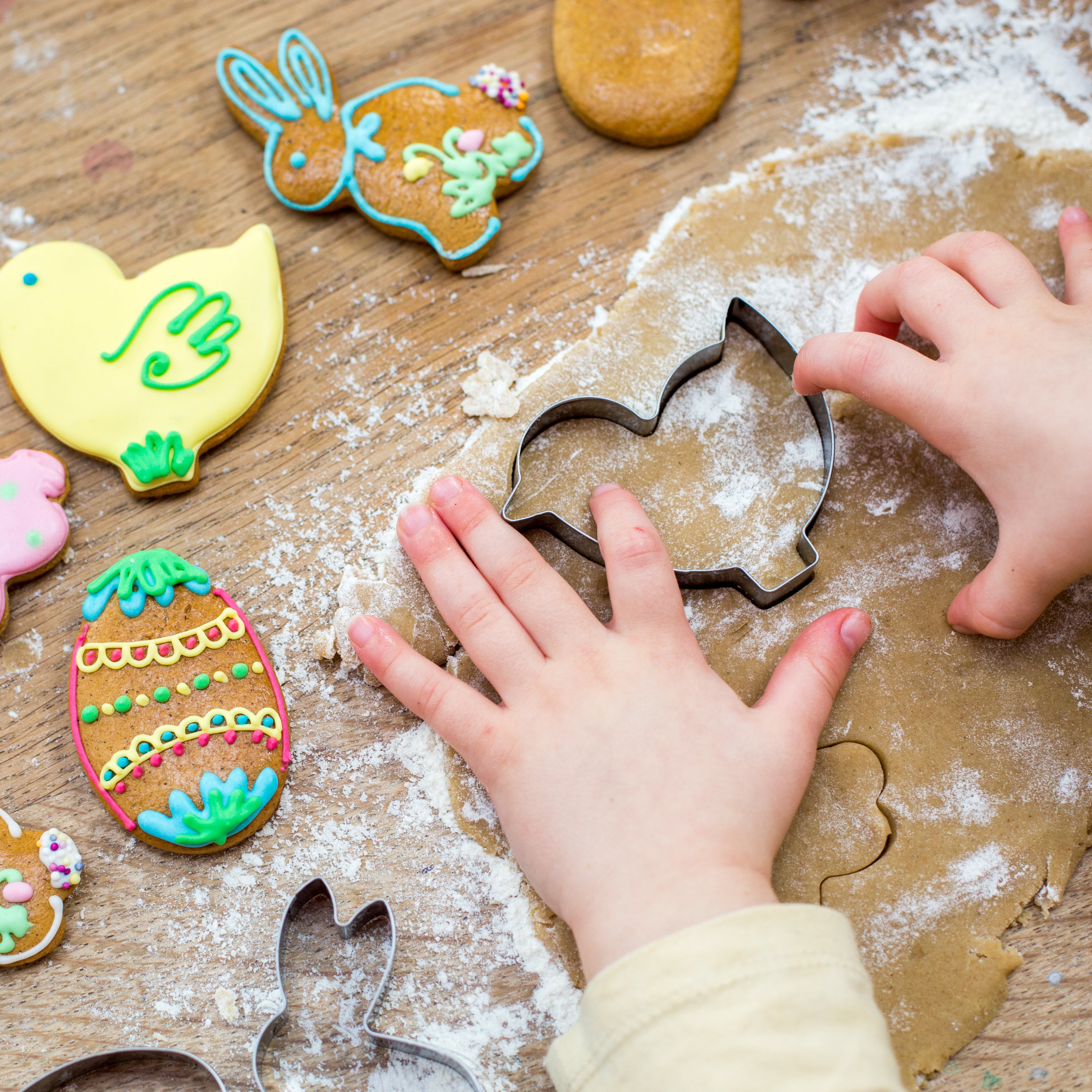 Child's hands using bunny cookie cutter on dough, surrounded by colorful Easter cookies on a floured wooden surface.
