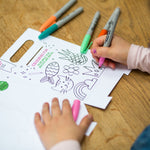 Child coloring a paper with markers, featuring a cat, pineapple, and rainbow on a wooden table.