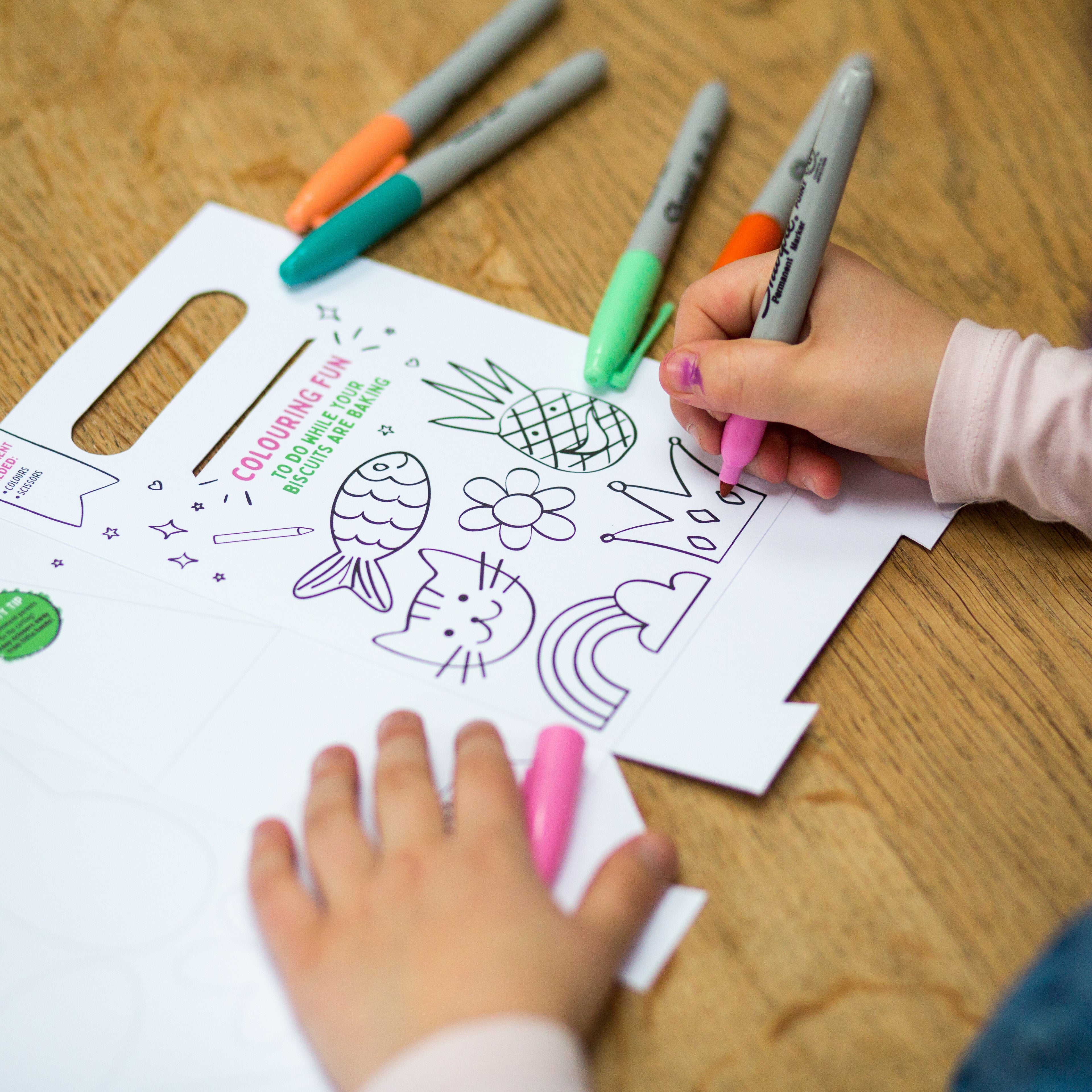 Child coloring a paper with markers, featuring a cat, pineapple, and rainbow on a wooden table.